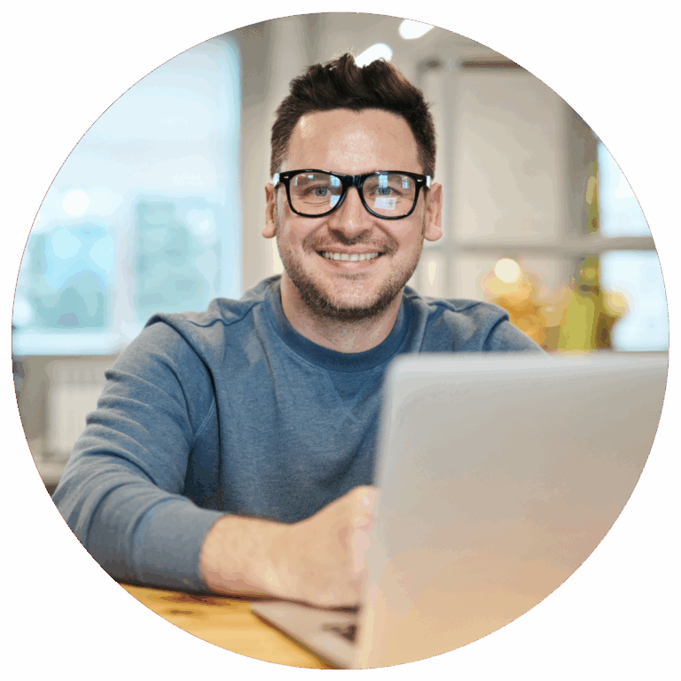 homme devant un ordinateur portable, sans un bureau d'entreprise avec un pull bleu et des lunette rectangulaire qui sourie.