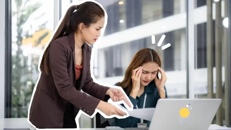 Une salariée stressée au bureau se tient la tête tandis qu’une collègue lui parle avec insistance, illustrant une situation de tension ou de conflit au travail.
