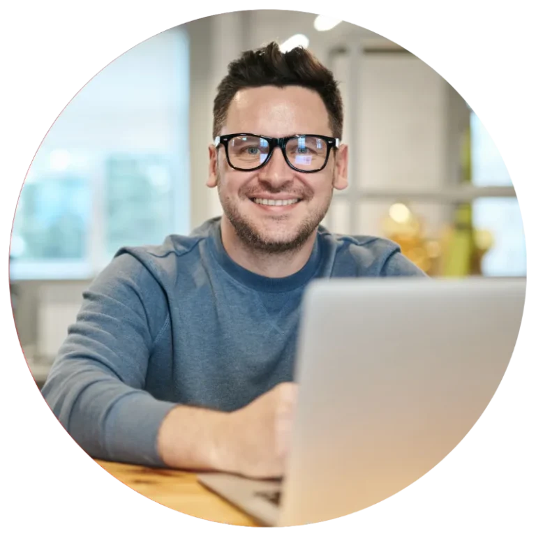 homme devant un ordinateur portable, sans un bureau d'entreprise avec un pull bleu et des lunette rectangulaire qui sourie.
